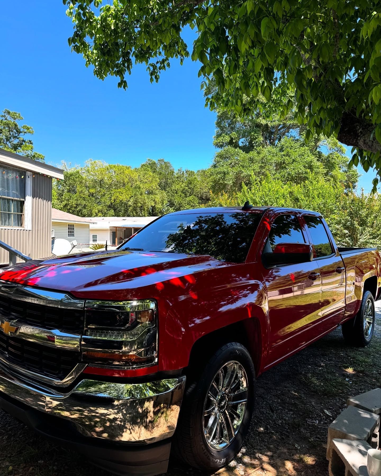 Red pickup truck parked in a shaded driveway beside a house, with trees and blue sky overhead