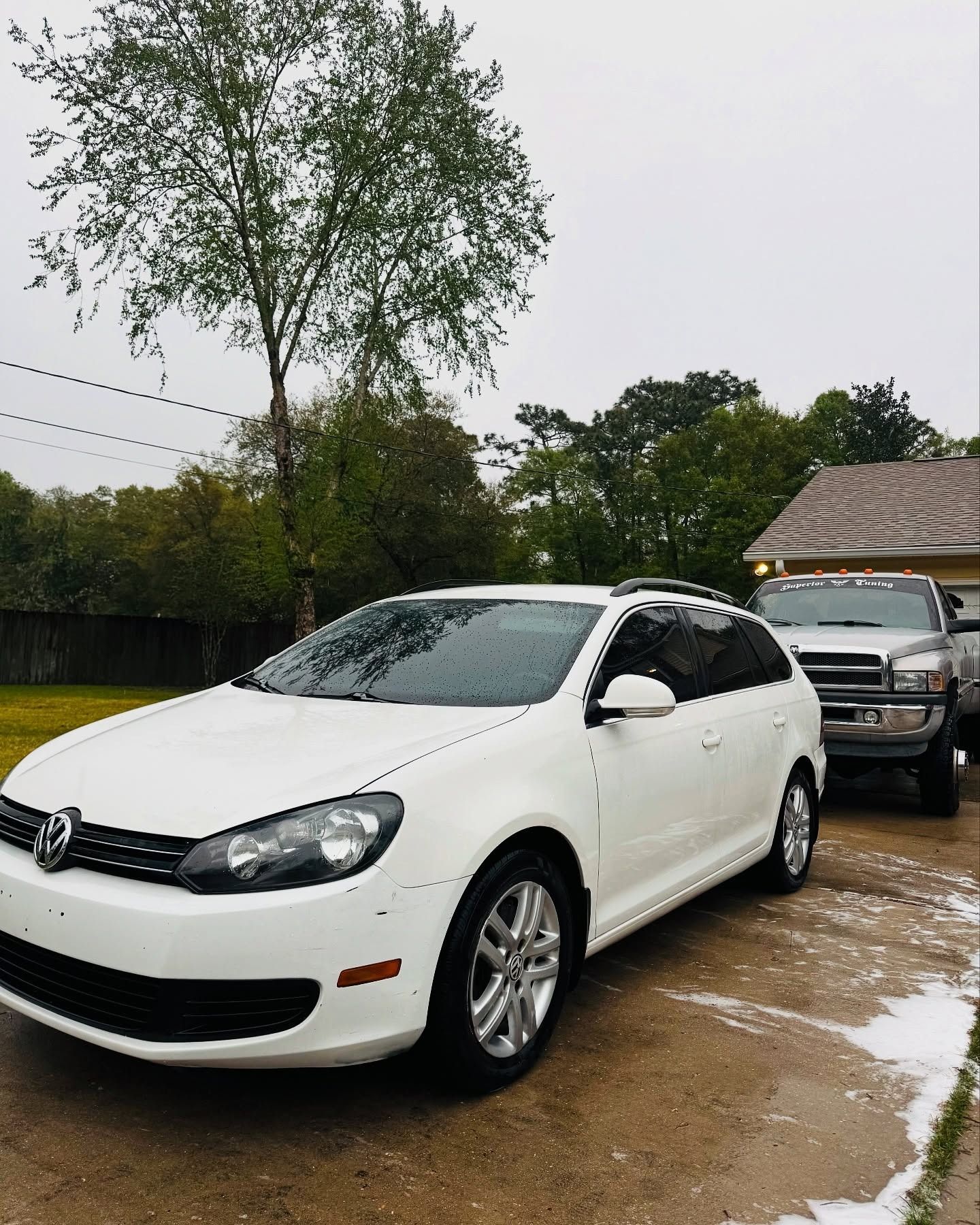 White hatchback parked on a wet driveway with a silver SUV behind it, trees and a house in the background