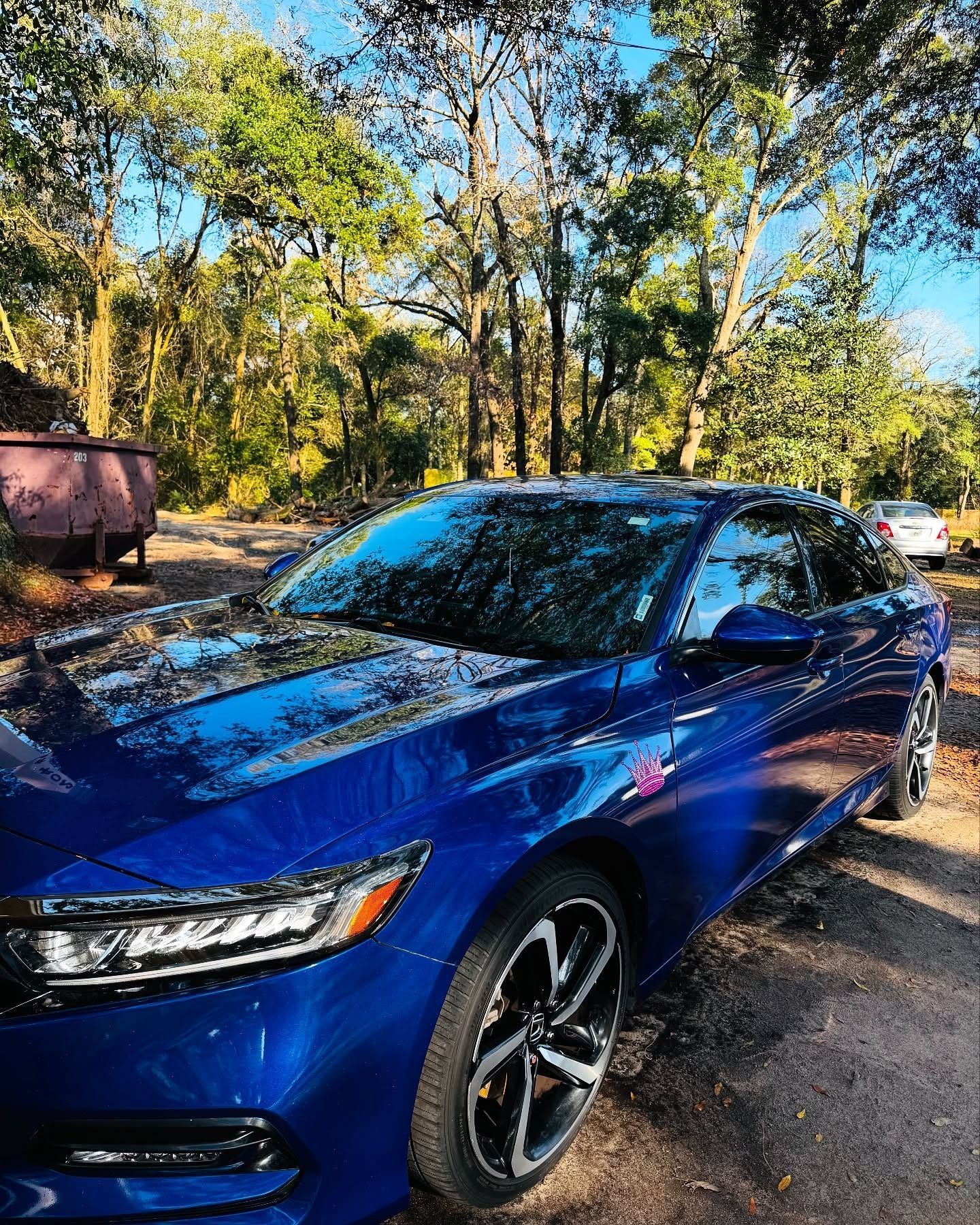 Blue sedan parked on a gravel road in a wooded area on a sunny day