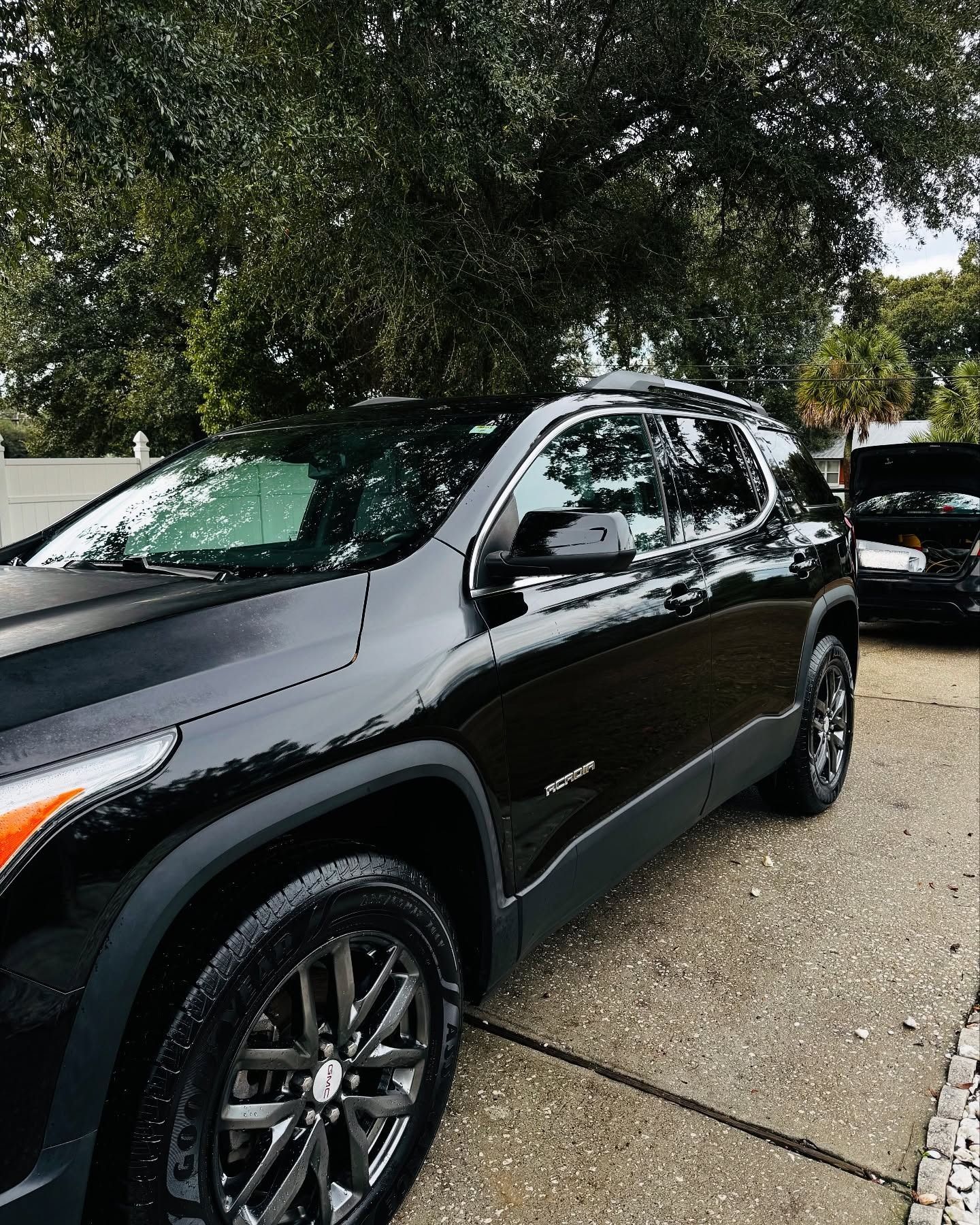 Black SUV parked on a gravel lot under trees, with another car behind it.