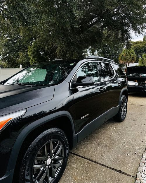 Black SUV parked on a gravel lot under trees, with another car behind it.
