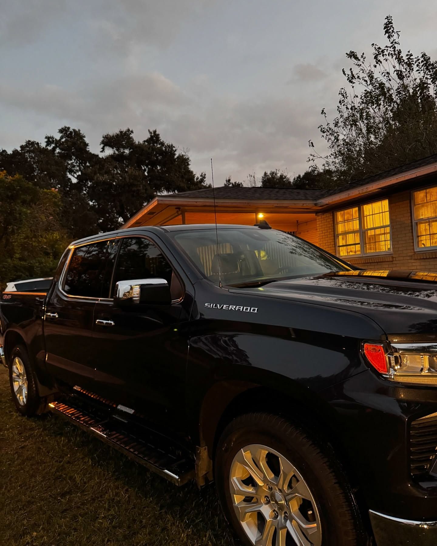 Black pickup truck parked beside a warmly lit cabin at dusk, with trees in the background