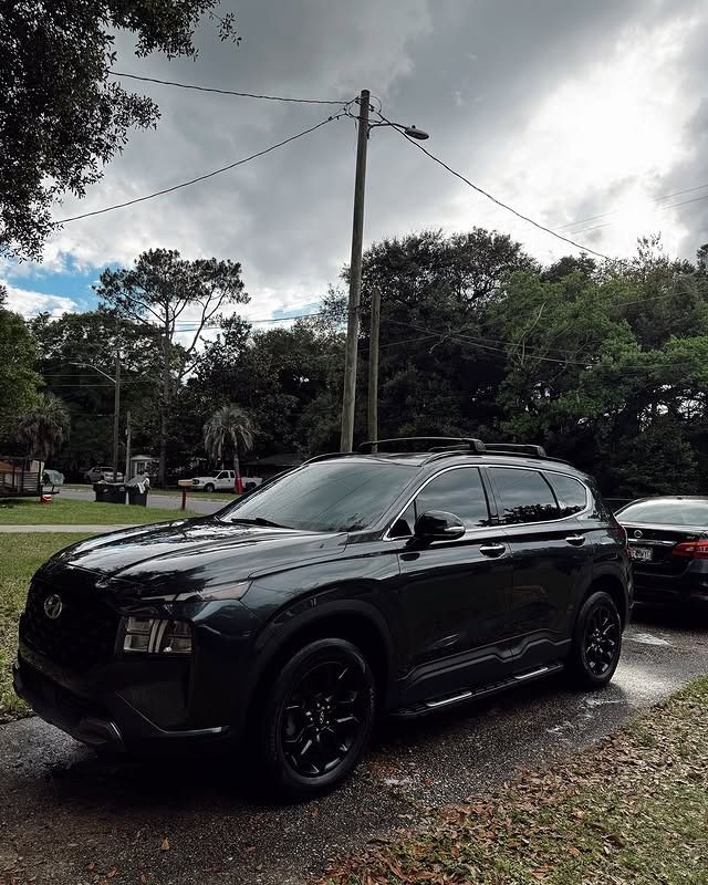 Black SUV parked on a wet roadside under cloudy skies, with trees and power lines in the background