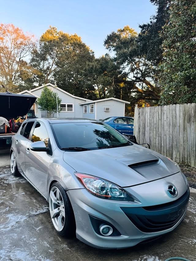 Silver hatchback car parked in a driveway beside a wooden fence and houses at sunset.