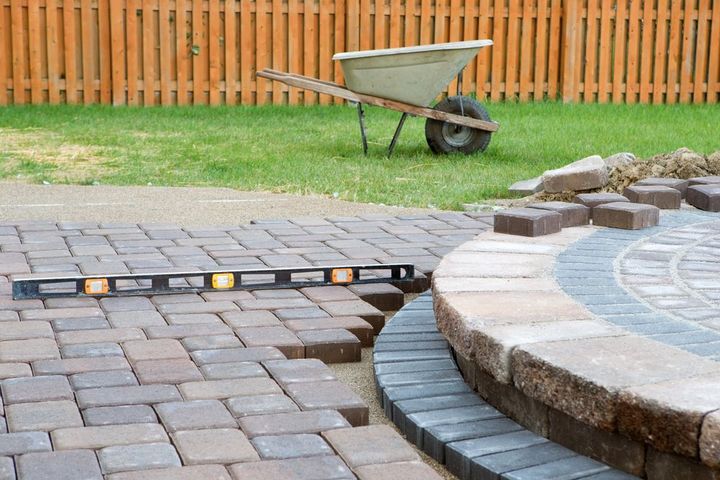 A level rests on unfinished patio pavers next to a raised stone fire pit in a backyard with a wheelbarrow and fence.