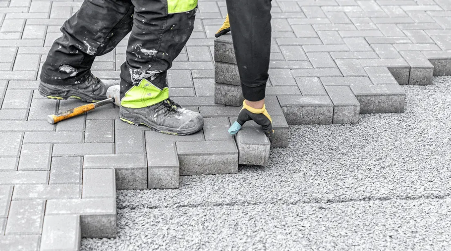A worker in work pants and gloves lays rectangular gray paving stones on a gravel base.