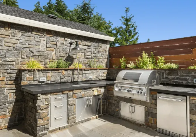 An outdoor kitchen featuring stone masonry, stainless steel grill, cabinets, sink, and wood fencing against a blue sky.