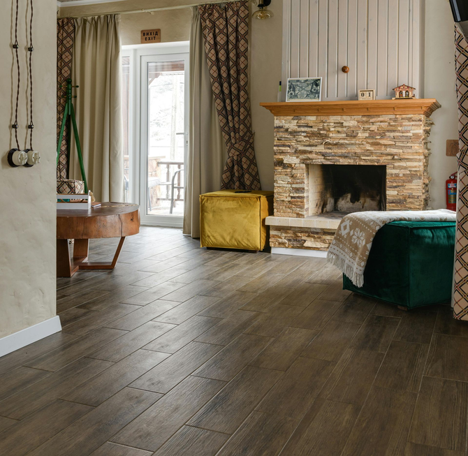 A cozy living room with rustic wooden floors, a stone fireplace, a yellow ottoman, and a green velvet armchair.