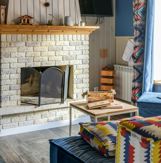 A cream brick fireplace with a metal screen, wood mantle, coffee table, and colorful patterned upholstery in a room.
