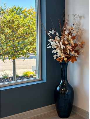 Tall dark vase with dried white and tan flowers beside a window in a blue-walled room