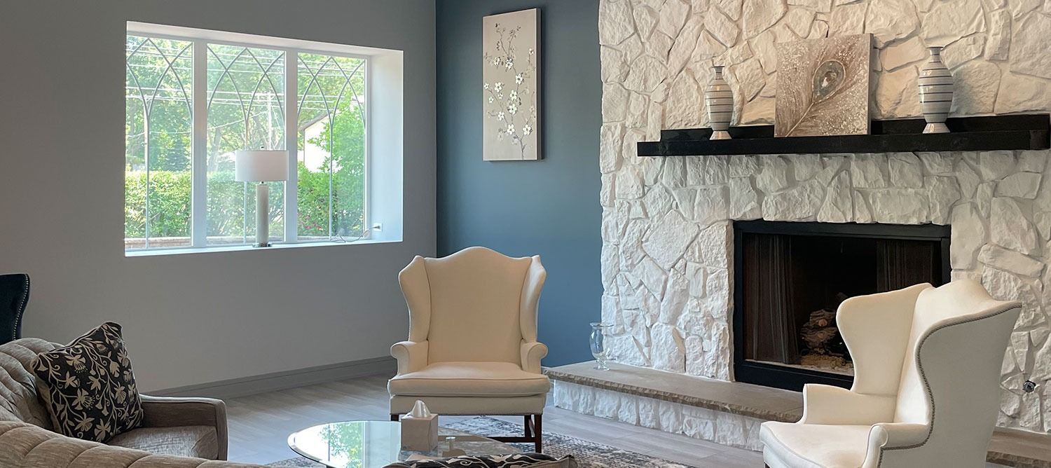 Bright living room with stone fireplace, white chairs, gray walls, and large window with greenery outside