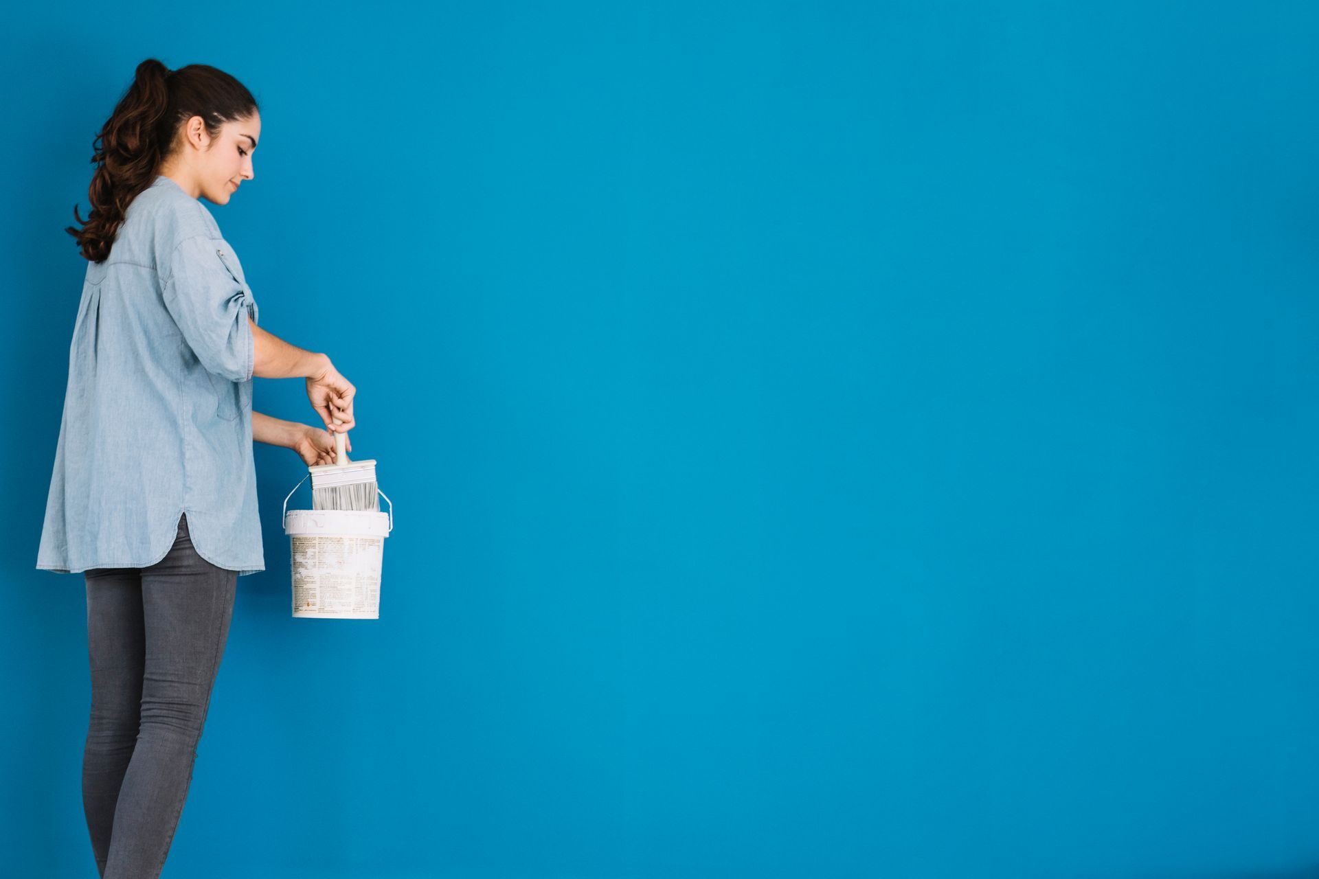 Woman dipping a paintbrush into a paint bucket in front of a blue wall.