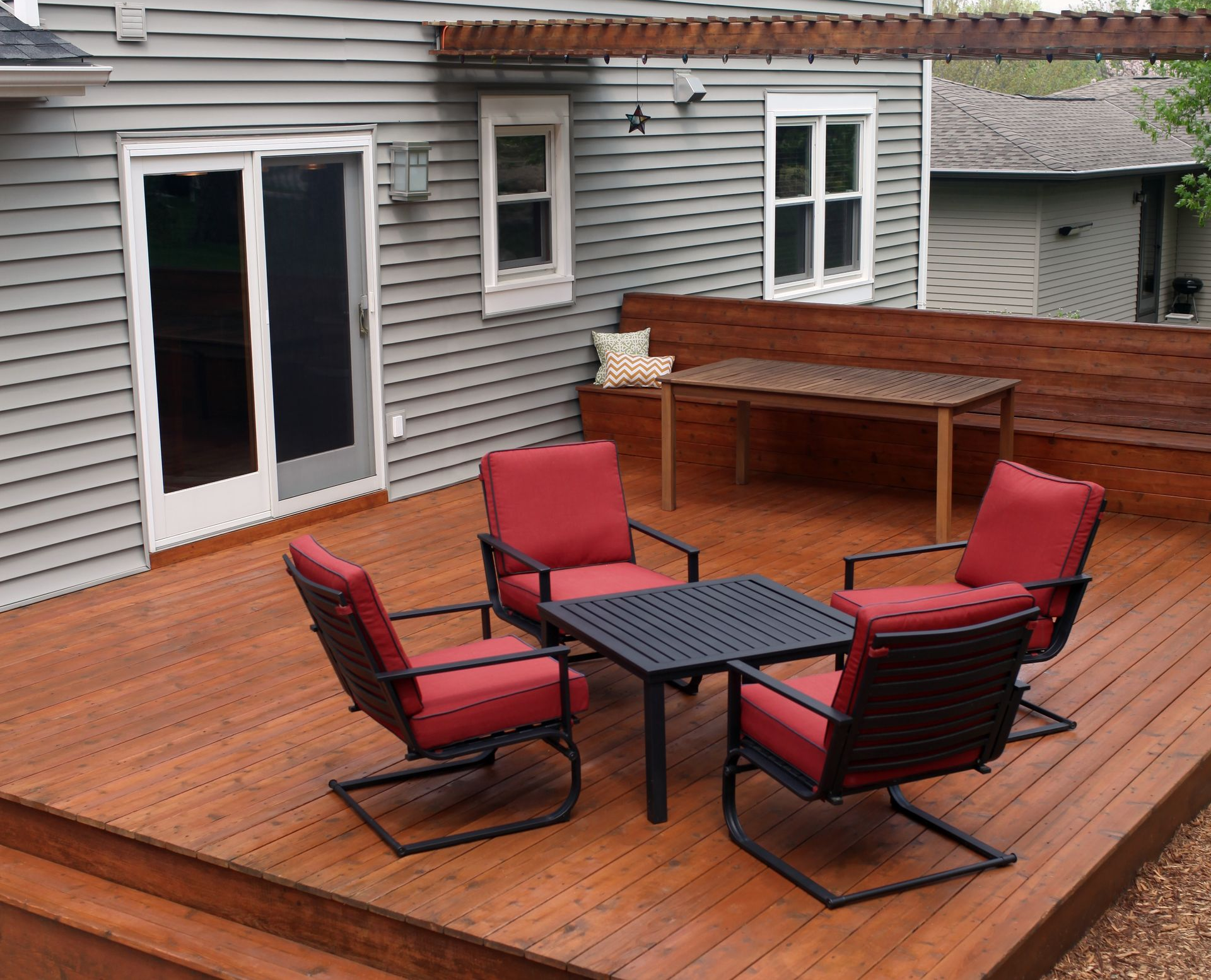 Wooden deck with red chairs around a square table. A long bench and dining table are in the background, next to a gray house.