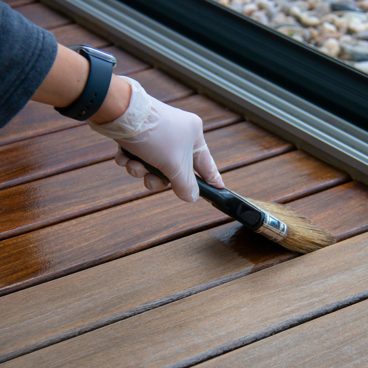 Gloved hand painting a wooden deck with a brush beside a sliding glass door