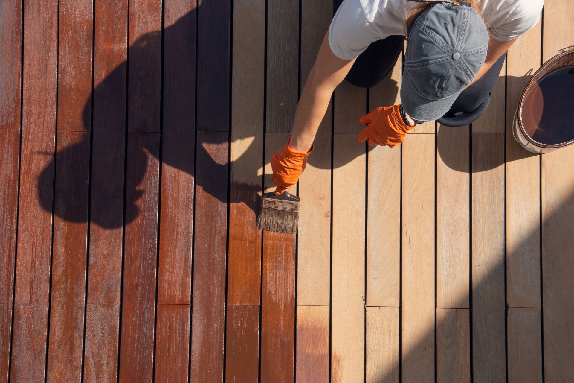 Person painting a wooden deck with a brush, showing a stained dark section next to unfinished boards.
