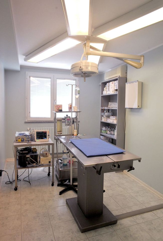 Sala visite mediche vuota con lettino, lampada a soffitto e armadietti vicino a una finestra.