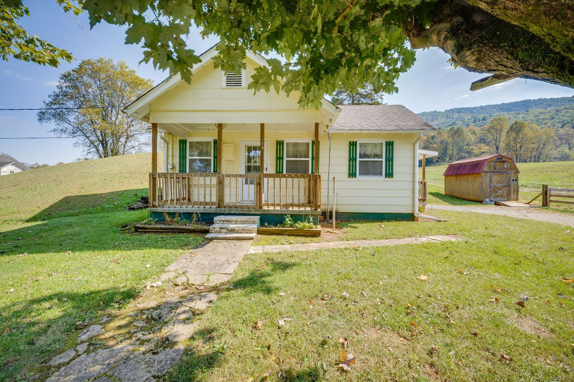 Small white farmhouse with a front porch on a grassy hill under a large tree.
