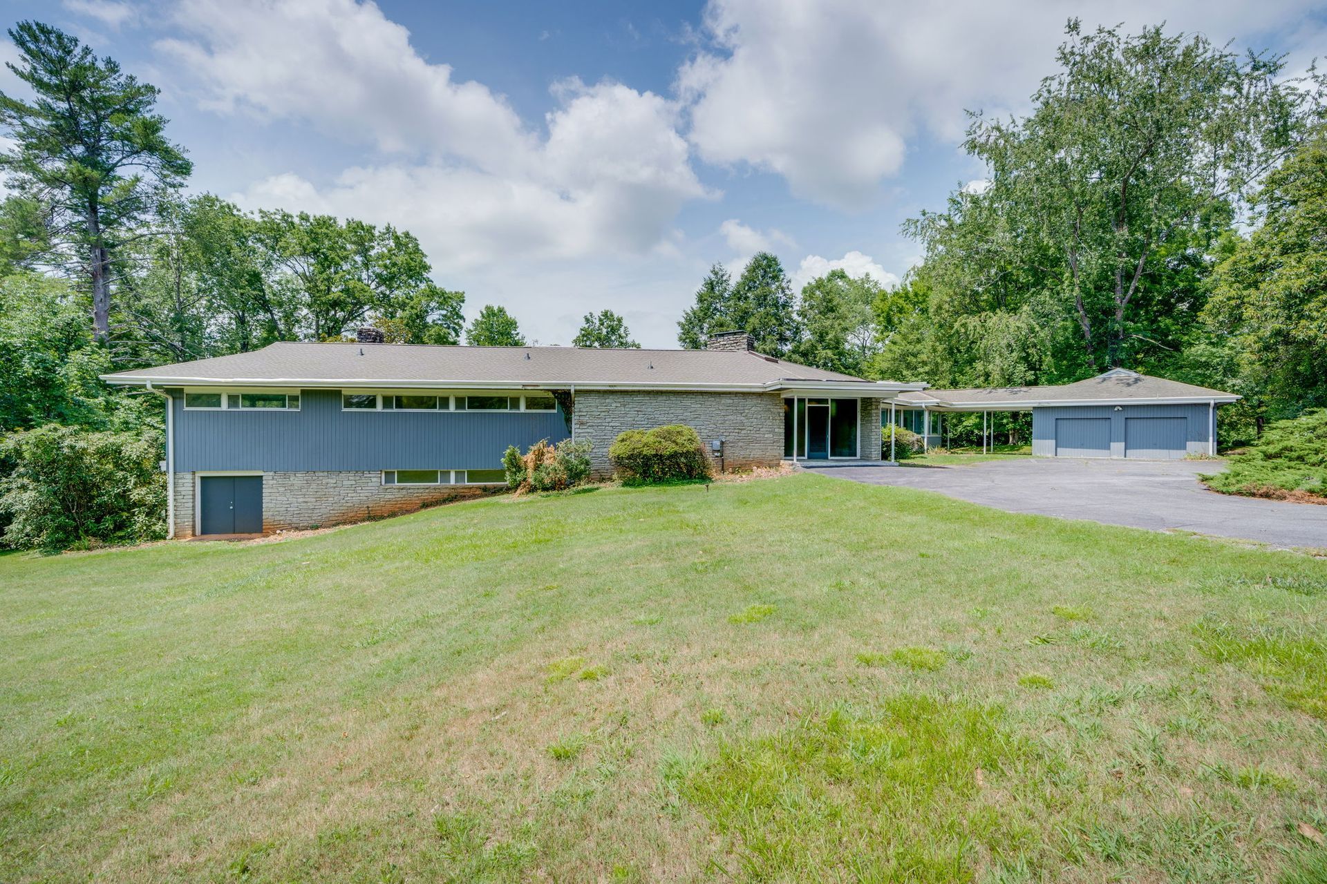 Single-story house with a large front lawn, trees, and a driveway under a partly cloudy sky