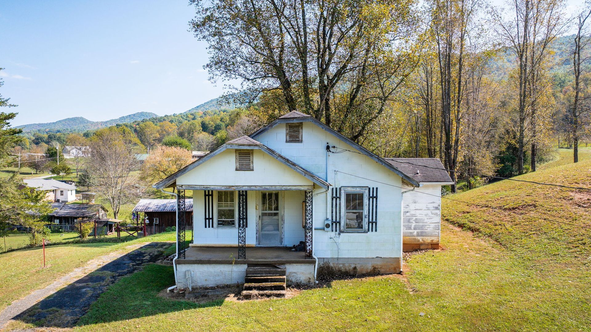 Small white house with a porch on a grassy hillside, surrounded by trees under a bright sky