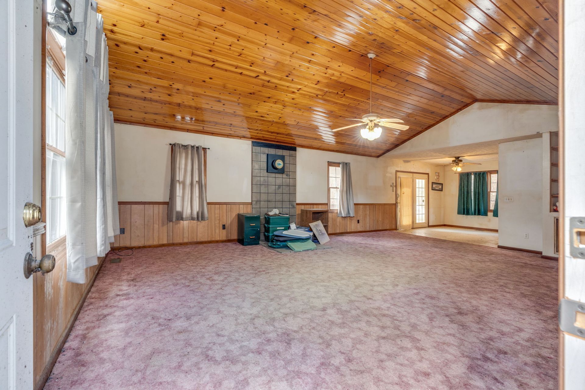 Spacious living room with wood ceiling, pink carpet, and a fireplace. Open doorway to another room.