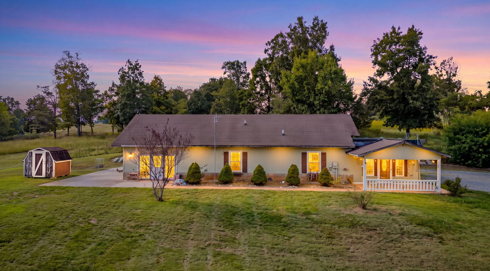 Cozy lit cottage with porch at dusk, surrounded by trees under a pink-purple sky.