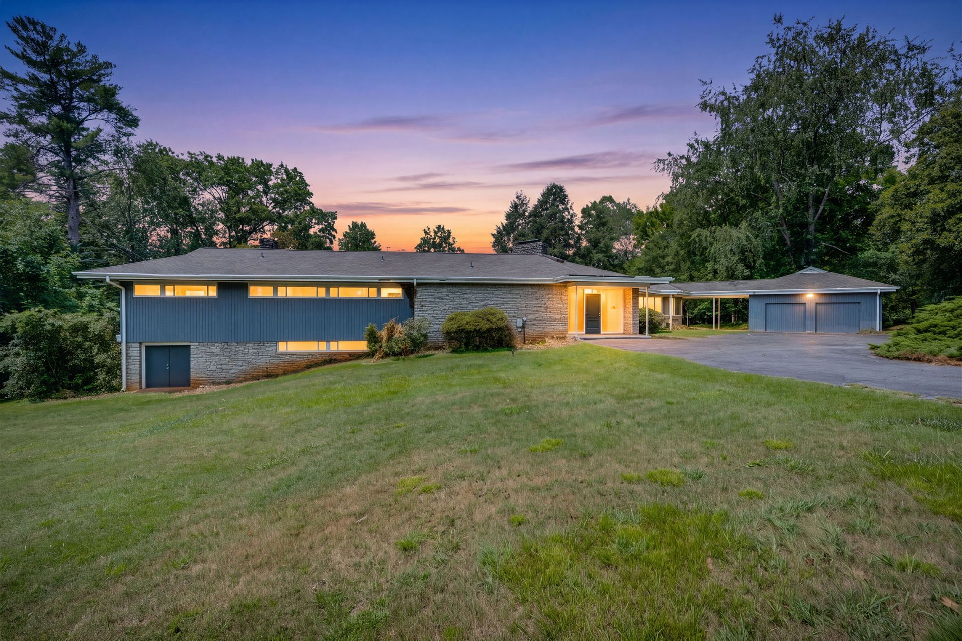 Modern single-story house at dusk with warm lights, a wide lawn, and trees surrounding the property.