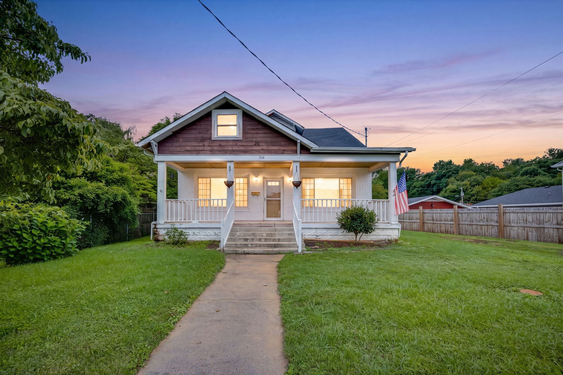 Small white house with a front porch and walkway at dusk, surrounded by a green lawn.