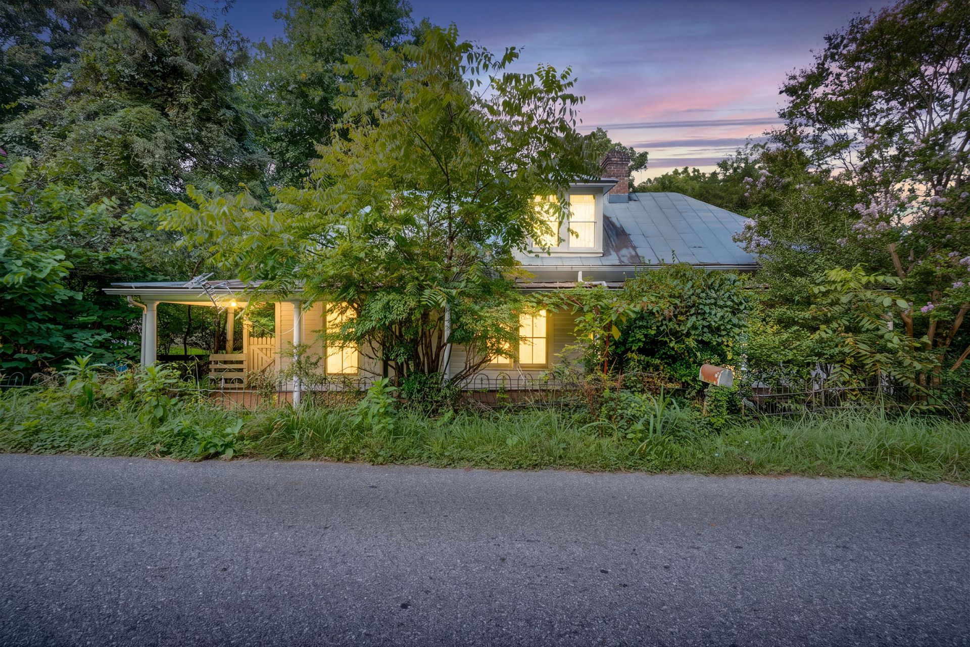 Small house with lit windows behind trees at dusk, viewed from a road.