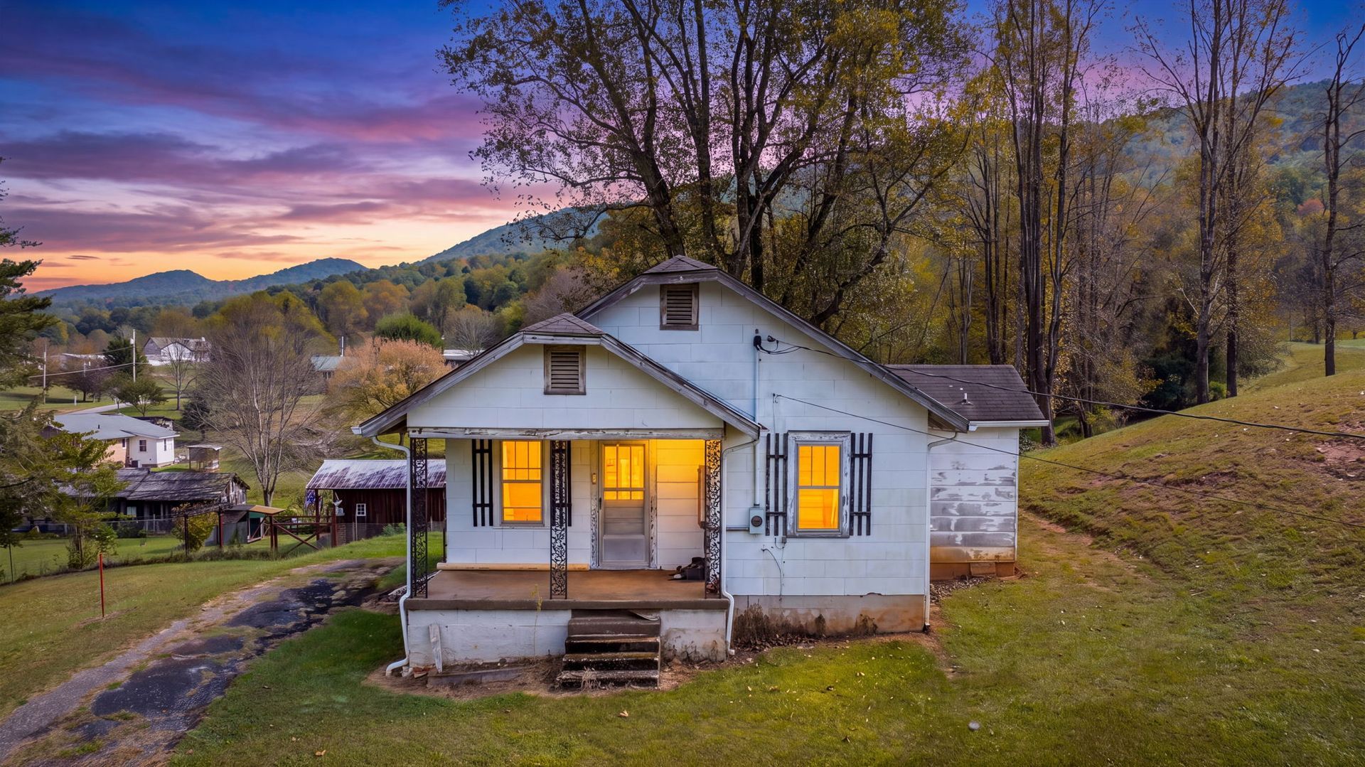 Small white house with lit windows on a hillside at dusk, under purple clouds and bare trees