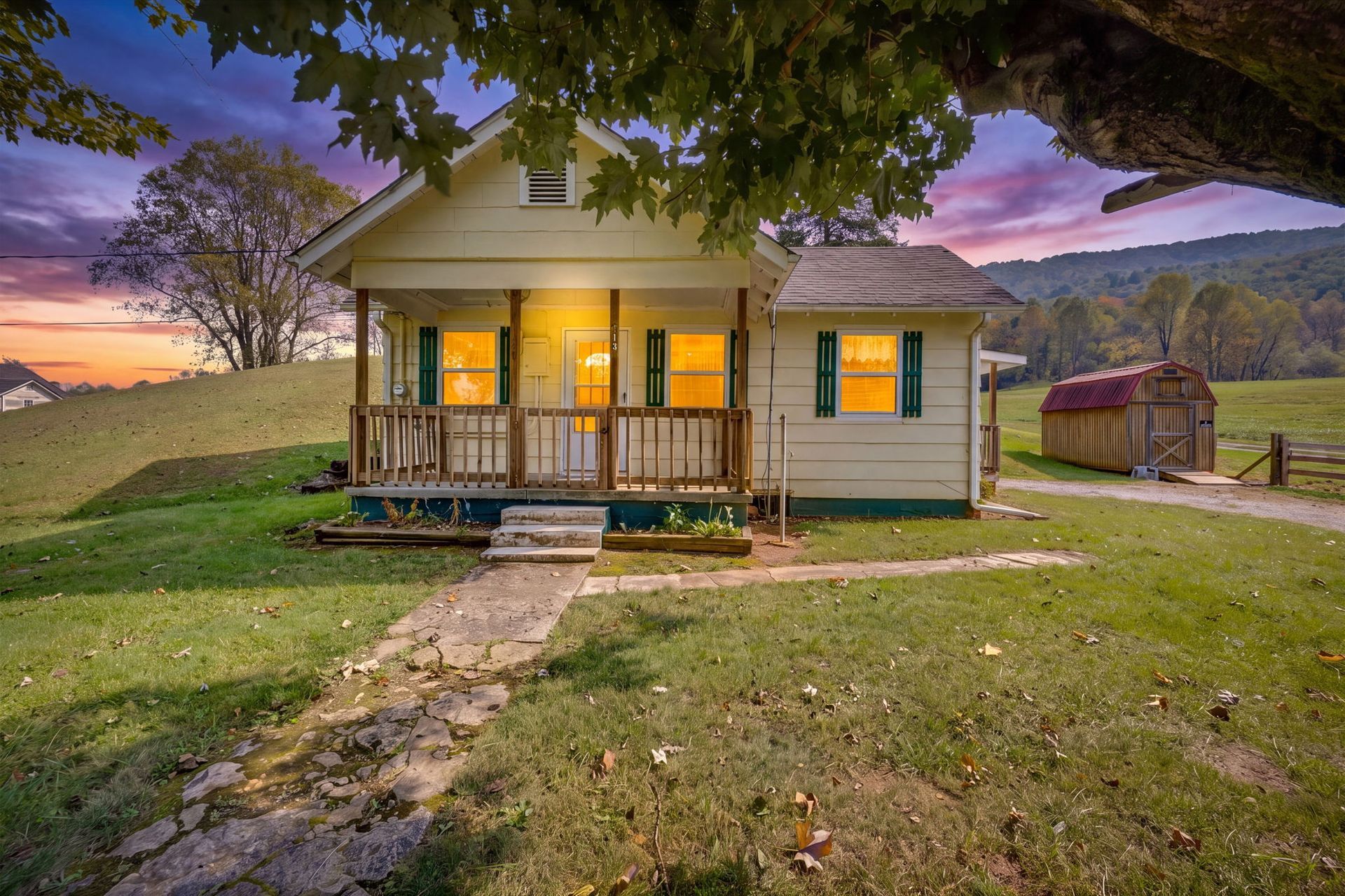 Small white farmhouse with lit porch at dusk, purple sky, grassy yard, and large tree branch framing the scene