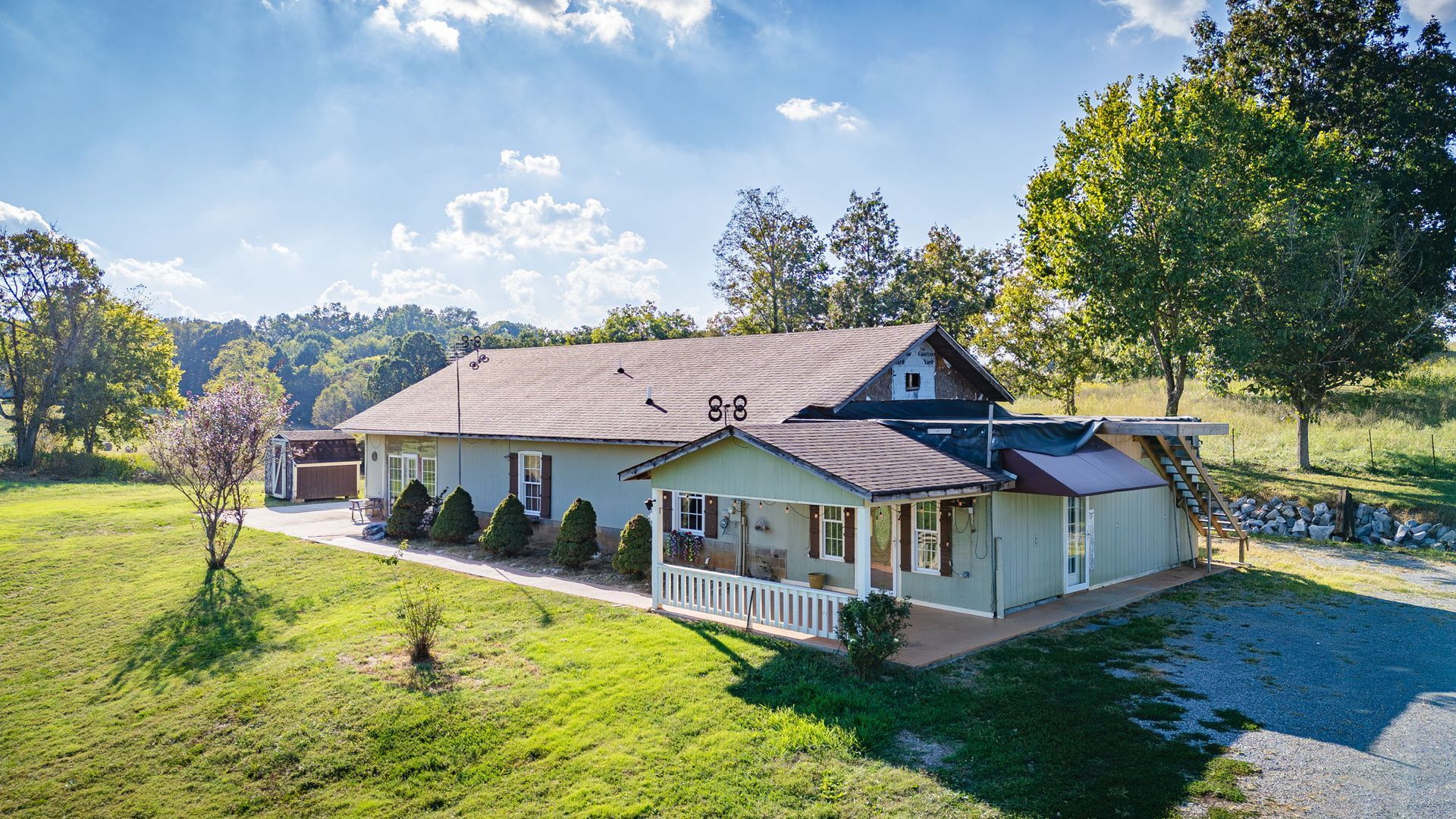 Countryside farmhouse with white porch, surrounded by green lawn and trees under a blue sky