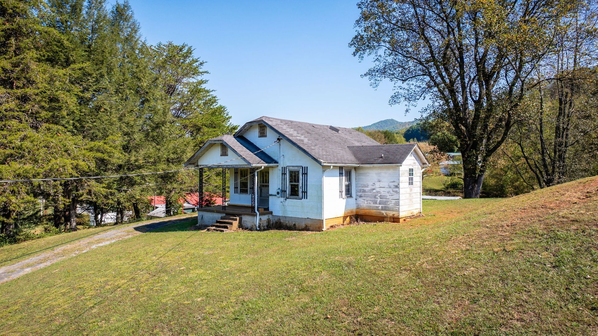Small white house with a porch on a grassy hillside, surrounded by trees under a clear blue sky