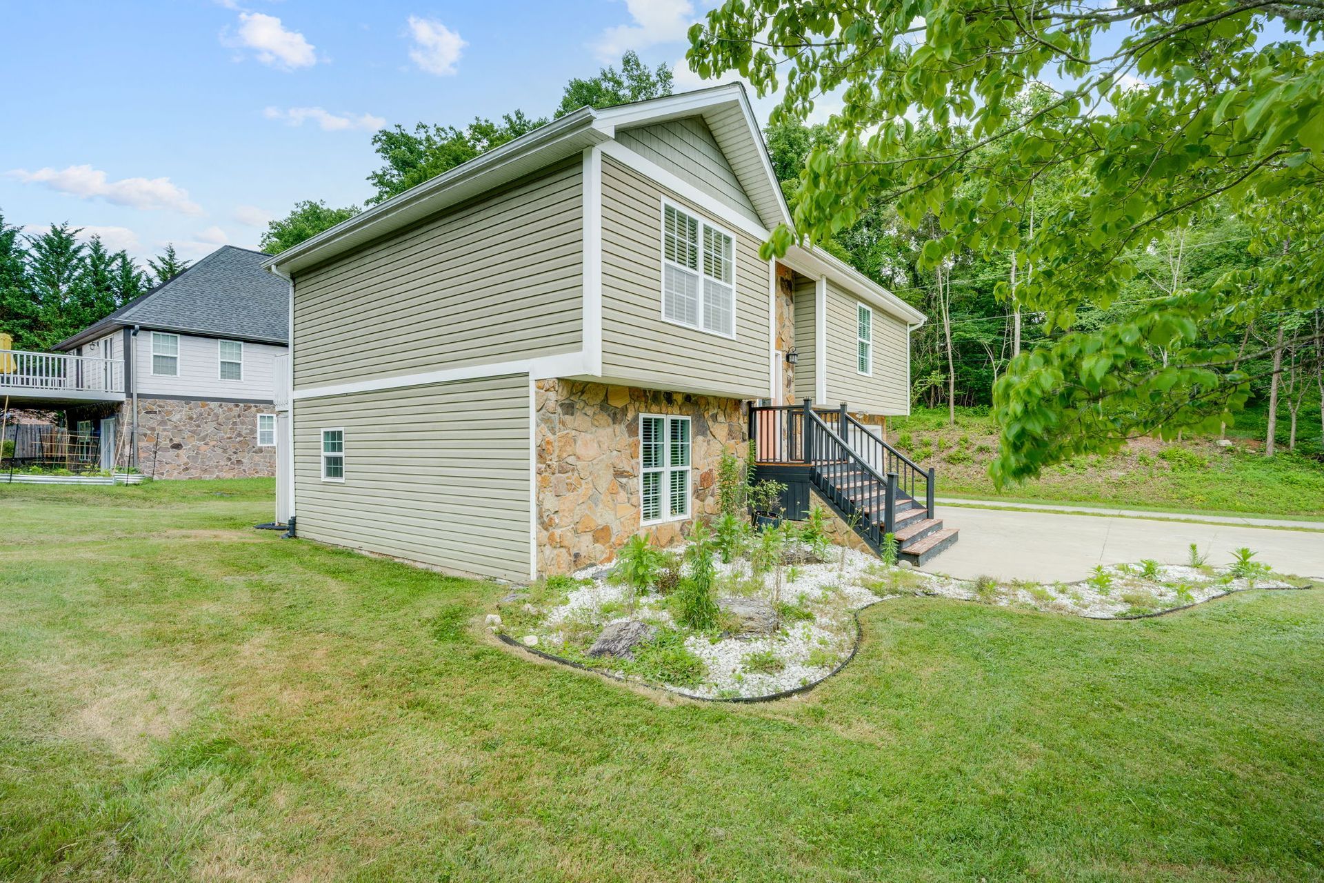 Two-story beige house with stone foundation, front stairs, and grassy yard surrounded by trees