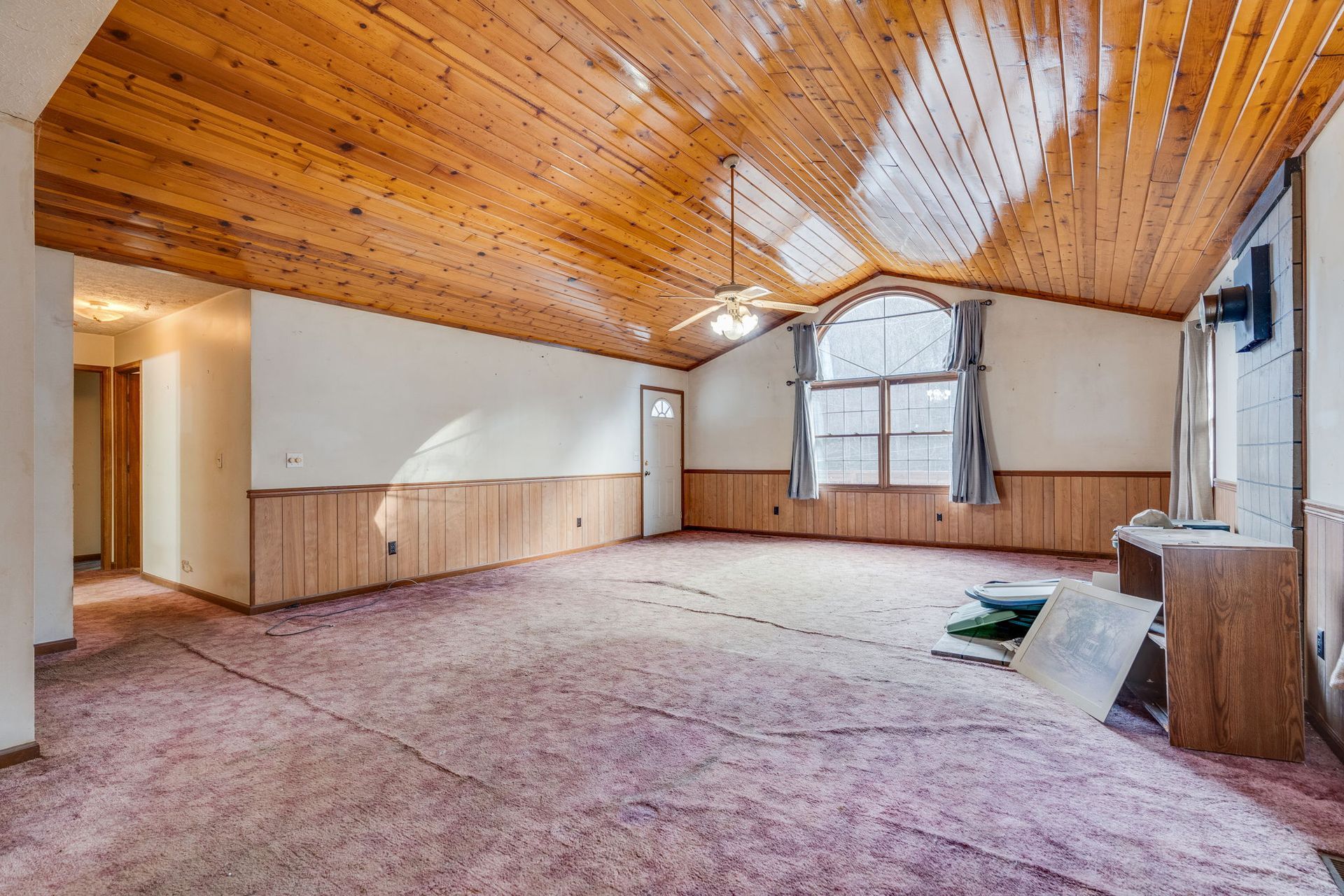 Spacious, empty room with wood panel ceiling and walls. Pink carpet, arched window.
