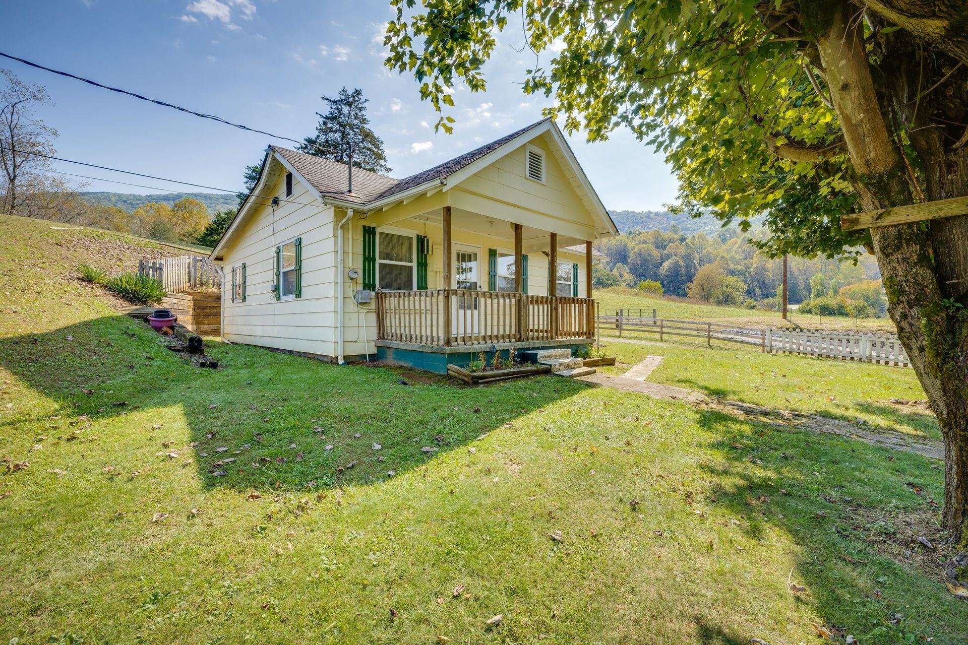 Small white farmhouse with a porch on a sunny grassy lot, framed by trees and rolling hills.