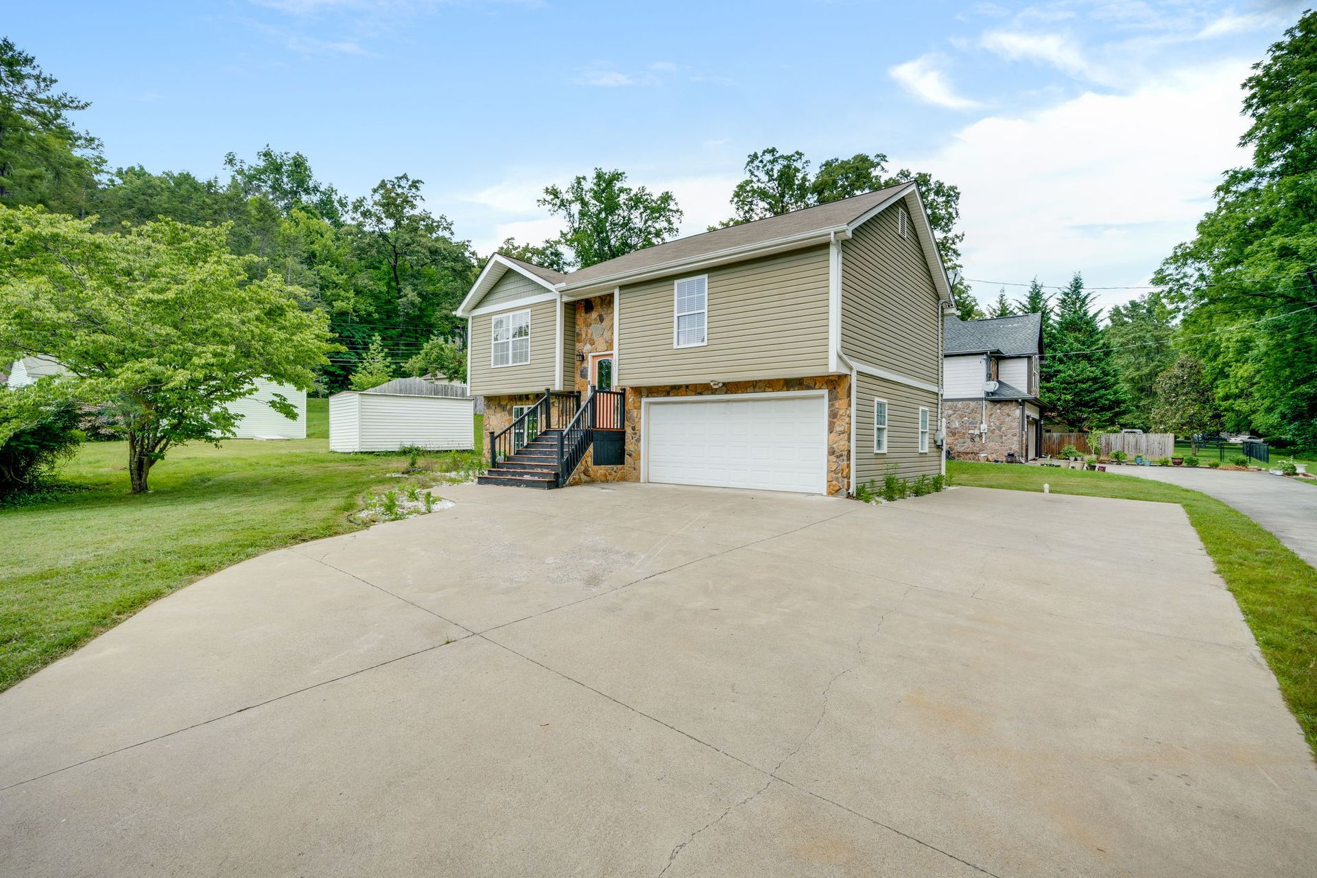 Two-story tan house with attached garage and large concrete driveway surrounded by green trees and lawn