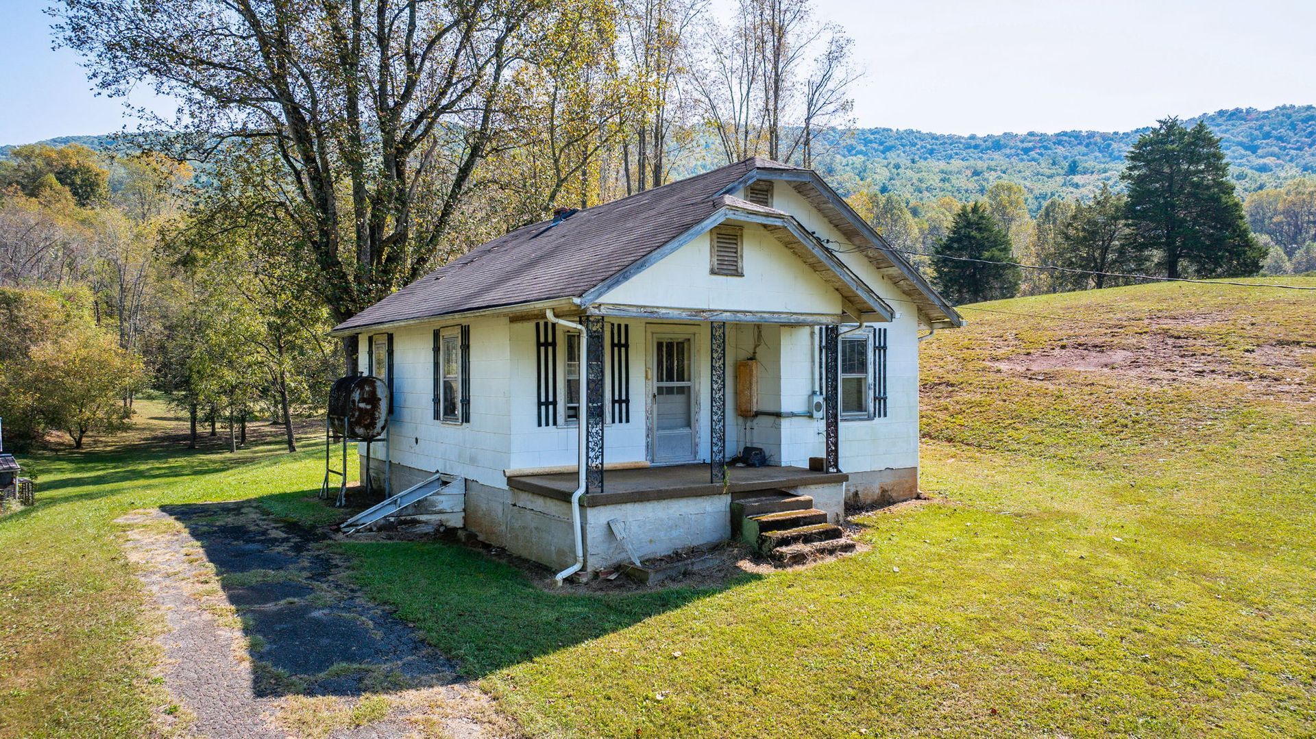Small white cottage with a porch and dark roof in a sunny rural field with trees and hills
