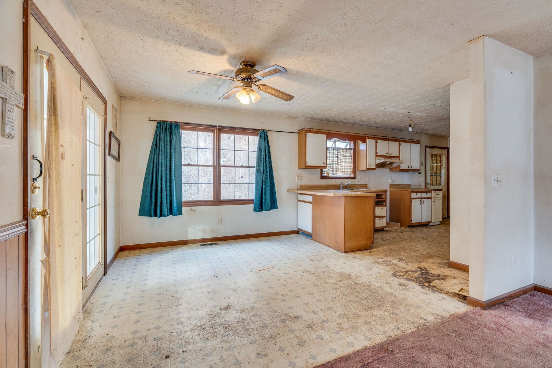 Interior of a home with open kitchen, dirty carpet, and teal curtains.