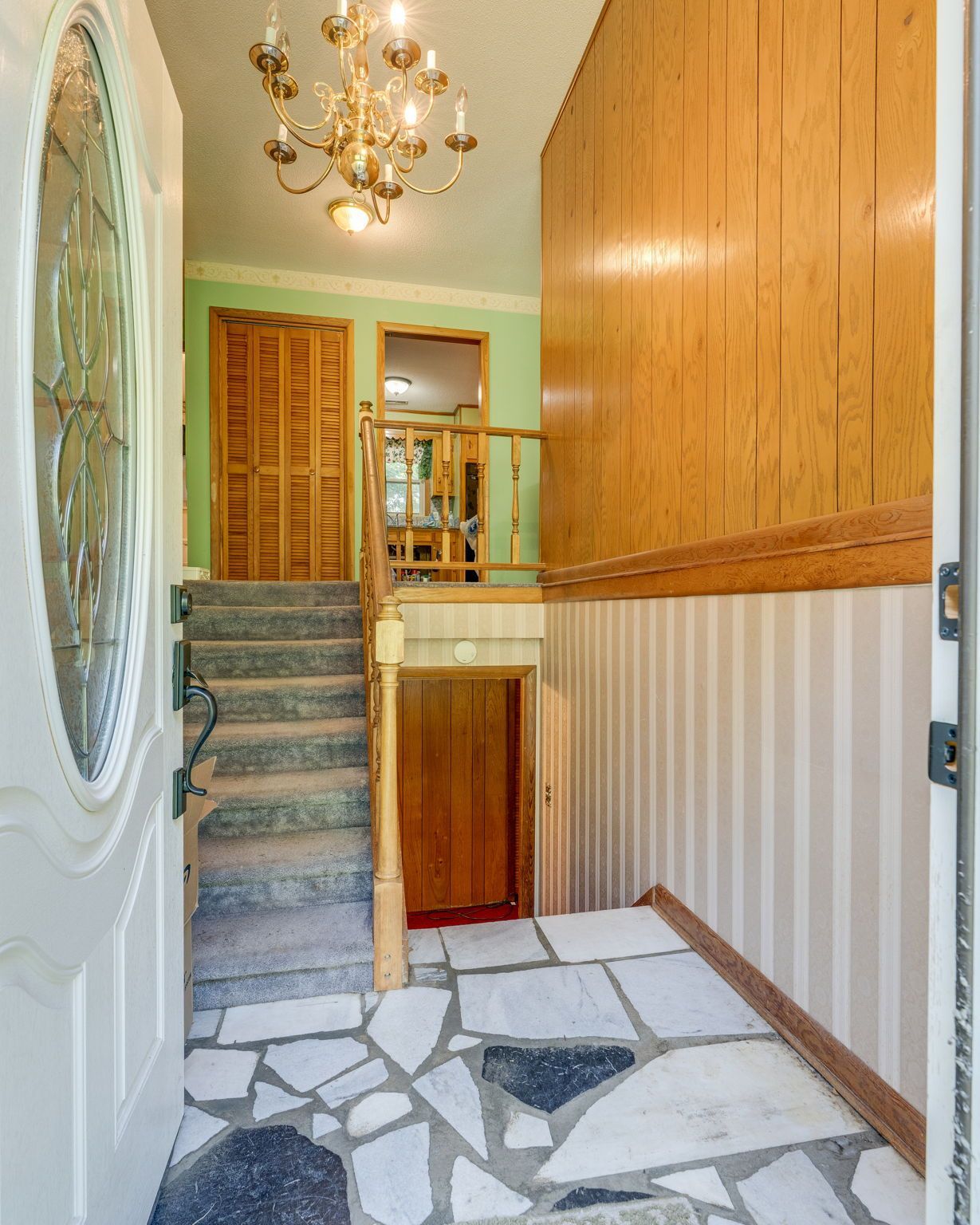Narrow hallway with white door, patterned tile floor, stairs, wood paneling, and chandelier lighting