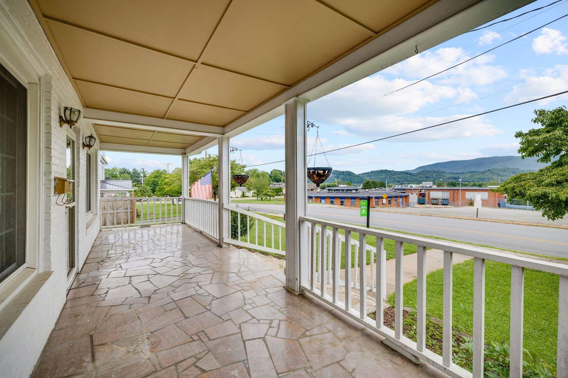 Covered porch with white railing overlooking a grassy yard, road, and distant hills under a partly cloudy sky