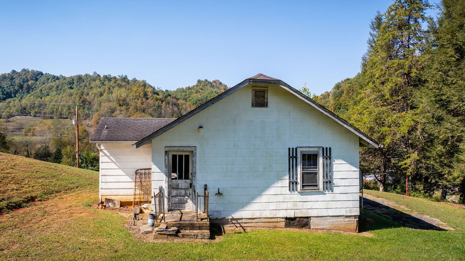 Small white house with a front porch in a grassy rural setting, surrounded by trees and hills
