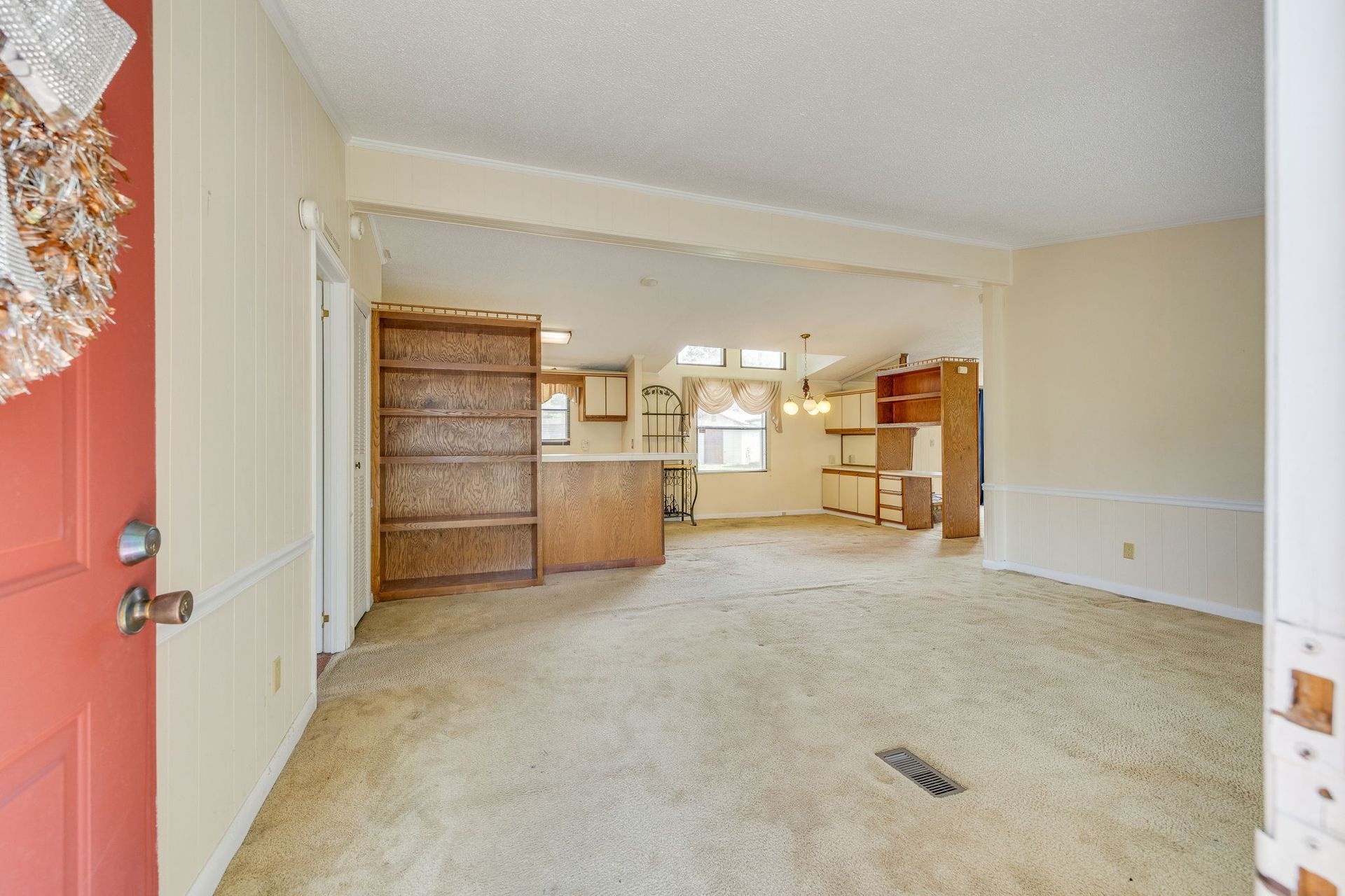Empty beige living room with carpet, wood cabinets, and a glimpse of kitchen area