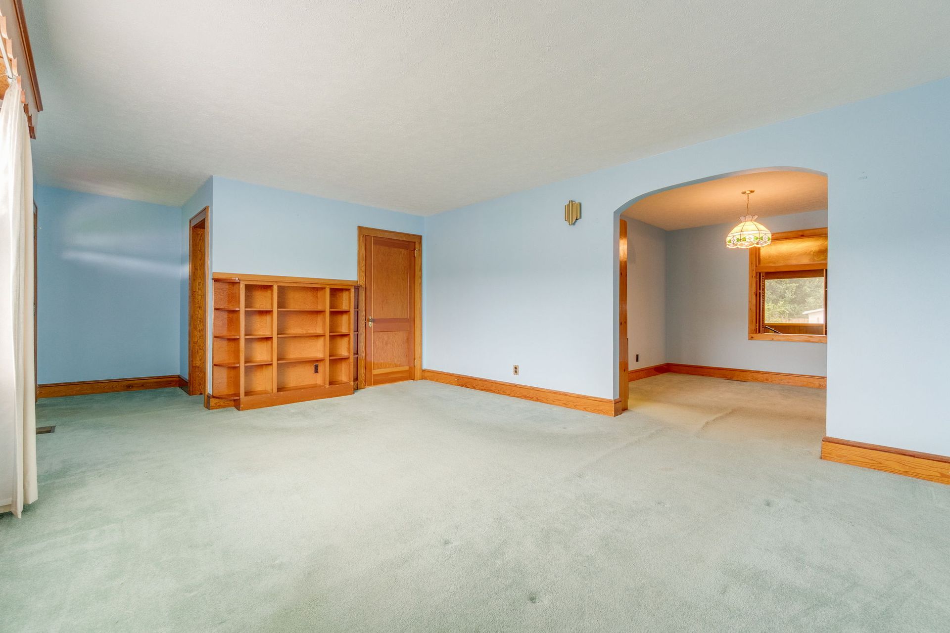 Empty carpeted living room with light blue walls, wooden trim, and arched doorway to a dining area