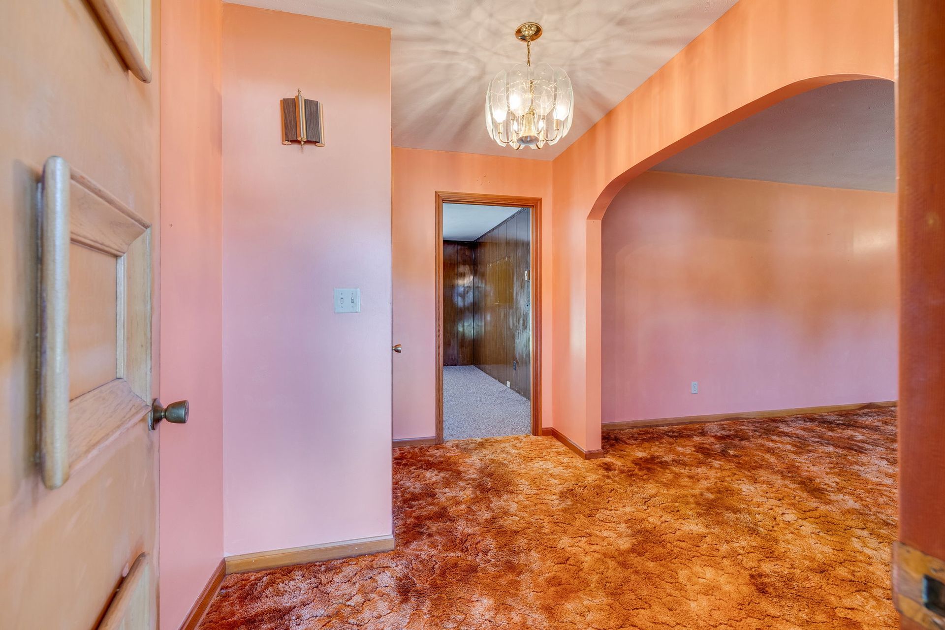 Empty pink-walled room with arched doorway, chandelier, and brown patterned carpet