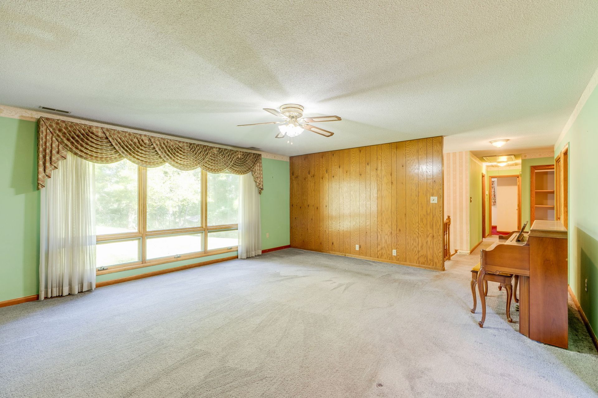 Empty living room with beige carpet, wood-paneled wall, large window, ceiling fan, and small desk by the doorway