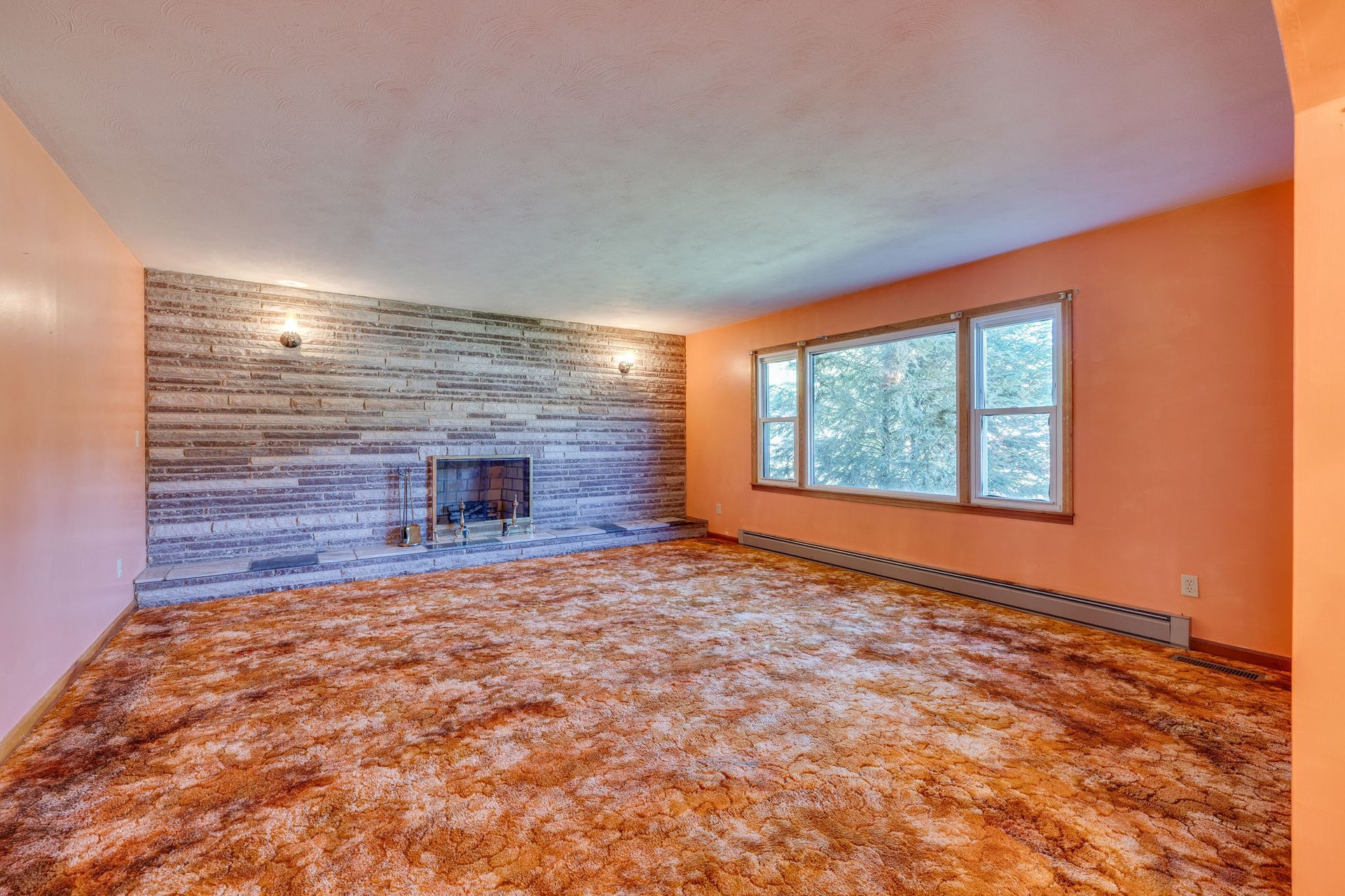 Empty living room with stone fireplace, orange carpet, and large windows on the right