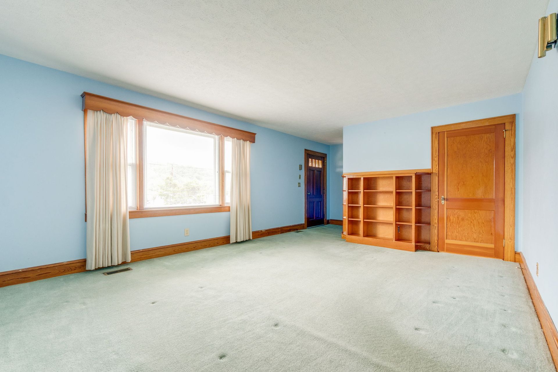 Empty carpeted living room with large window, built-in shelves, and blue front door