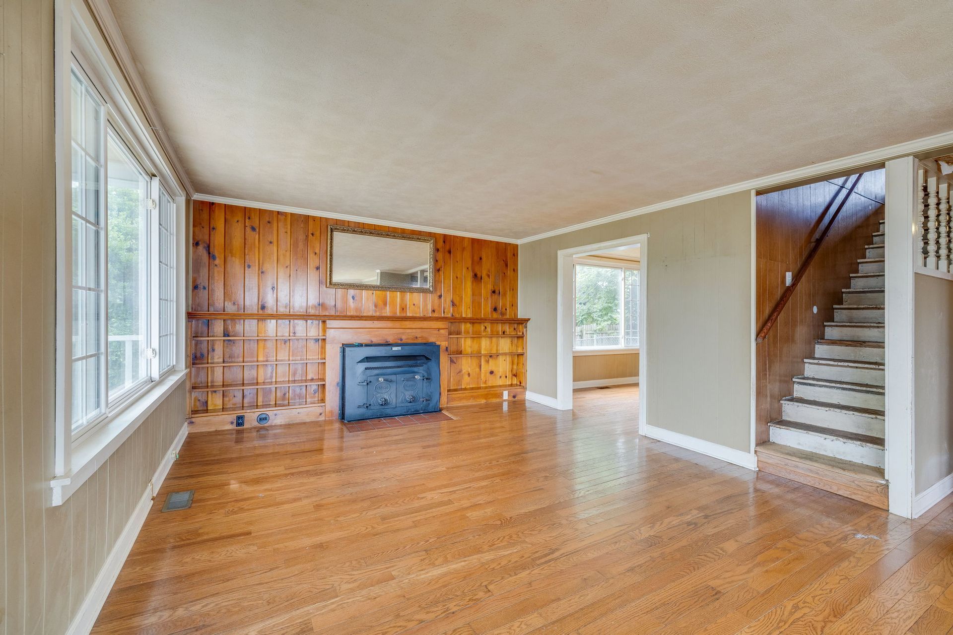 Empty living room with hardwood floors, wood-paneled accent wall, fireplace, and staircase at right