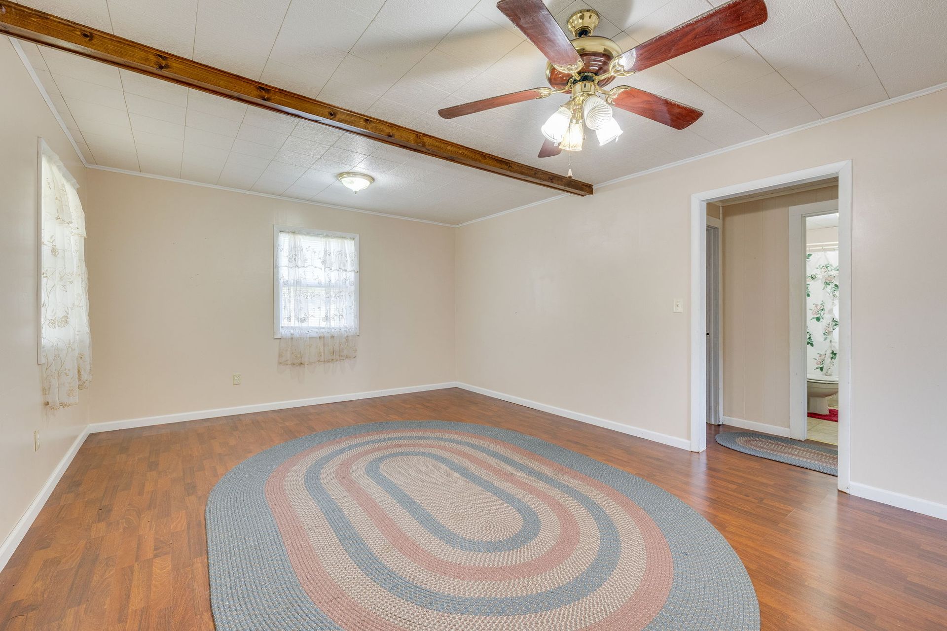 Empty beige bedroom with hardwood floor, ceiling fan, small window, and oval rug