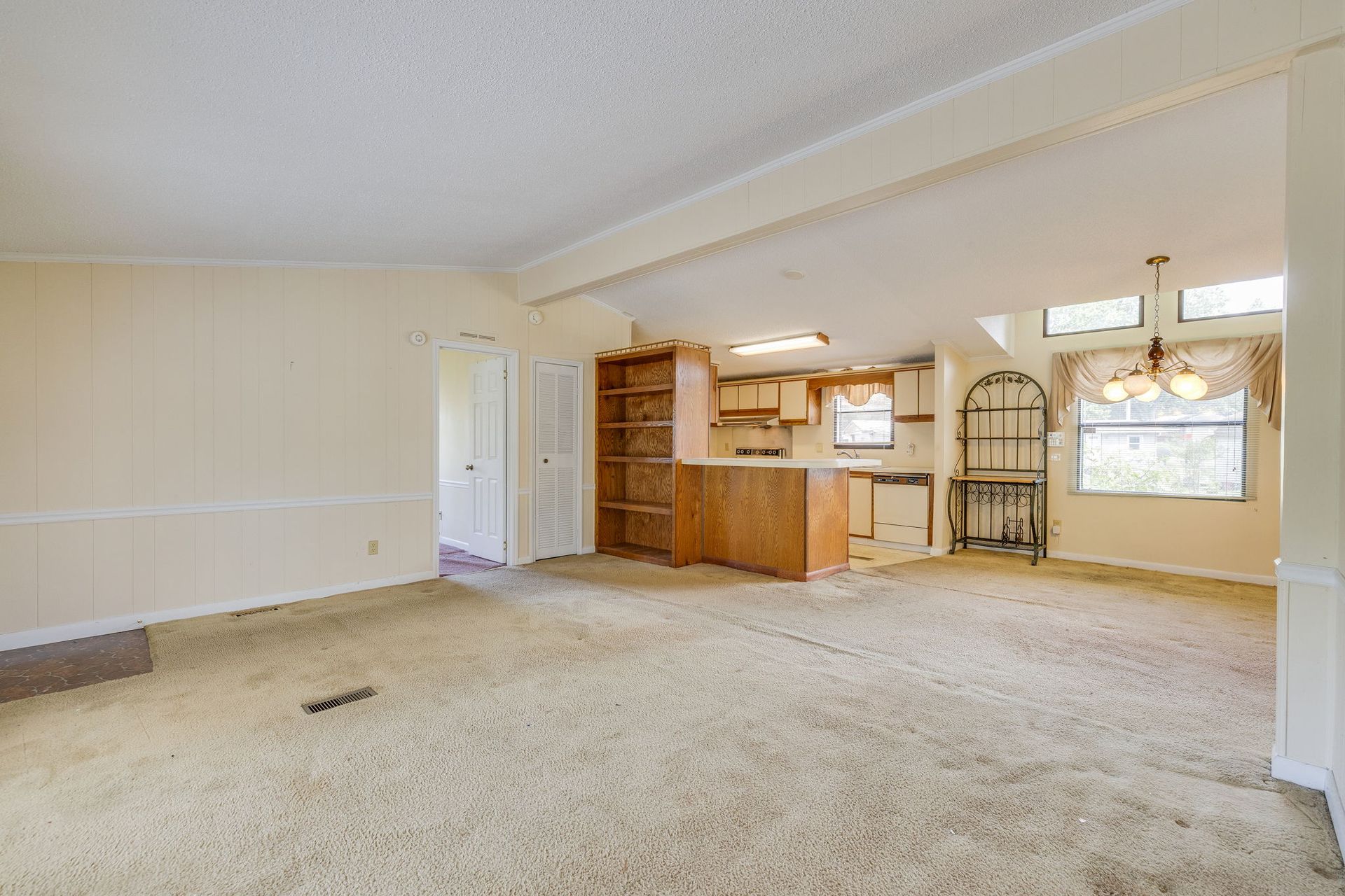 Empty beige living room with carpet, wood kitchen pass-through, and arched window.